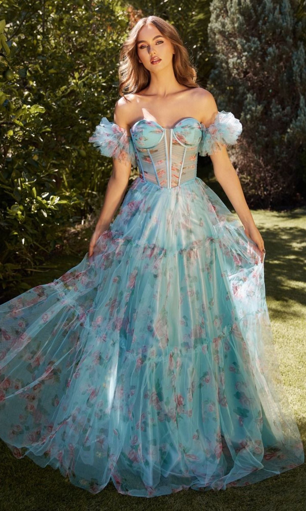 A woman stands outdoors in sunlight wearing the Puff-Sleeve Long Blue Print Prom Dress A1285, featuring a corset-style bodice and layered tulle skirt. She gently holds the skirt with both hands, showcasing its elegant flow.